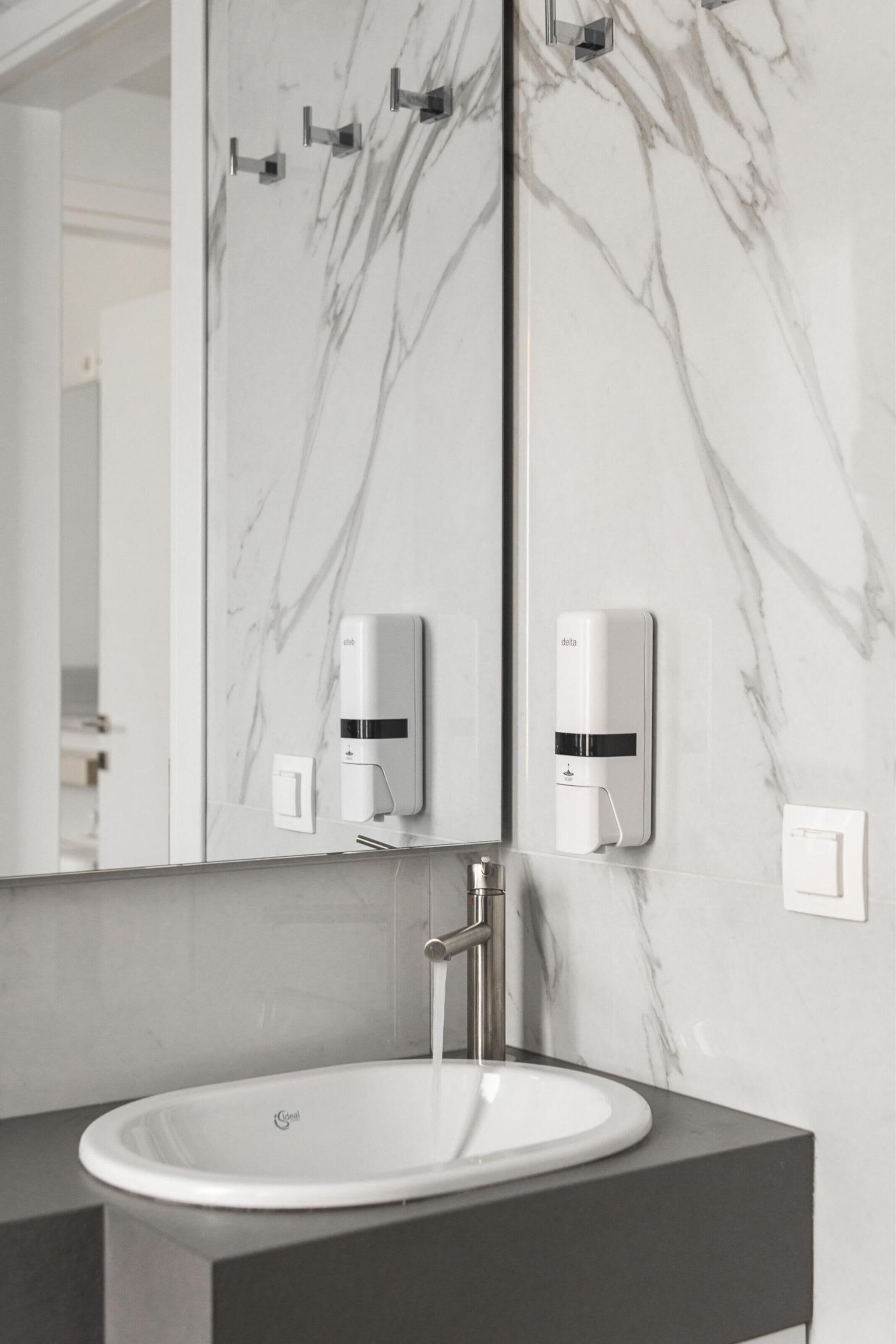 A bathroom with a marble wall, a white sink, and two soap dispensers.