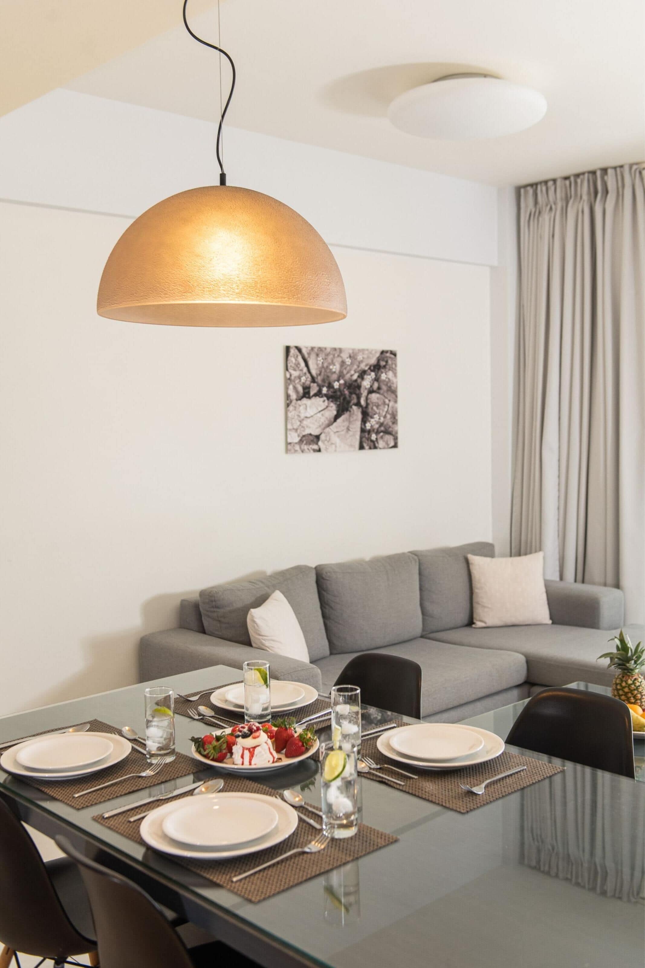 A dining area with a glass table, grey sofa, and a pendant light.