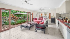 A spacious living area with a sofa, coffee table, and ceiling fans. A pool and lush greenery are visible through large windows. - Belle Escapes - Latania Villa Palm Cove (Palm Cove)