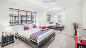A modern bedroom with a large bed, a ceiling fan, a window with blinds, a bedside table with a lamp, and a mirror. - Belle Escapes - Latania Villa Palm Cove (Palm Cove)