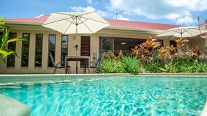 A swimming pool with a red-roofed building in the background, surrounded by plants and umbrellas. - Belle Escapes - Latania Villa Palm Cove (Palm Cove)