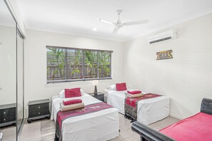 Une chambre à coucher propre et moderne, dotée de deux lits, d’un ventilateur de plafond, d’un climatiseur et d’une fenêtre avec des stores.