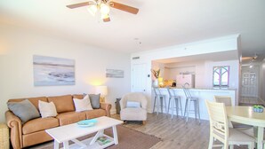 A living room with a sofa, coffee table, and chairs, a kitchen area with bar stools, and a ceiling fan. -  New 1BR/1BA Luxury Beachfront Vacation Rental! Sleeps 6! Near Pier Park! (Panama City Beach)