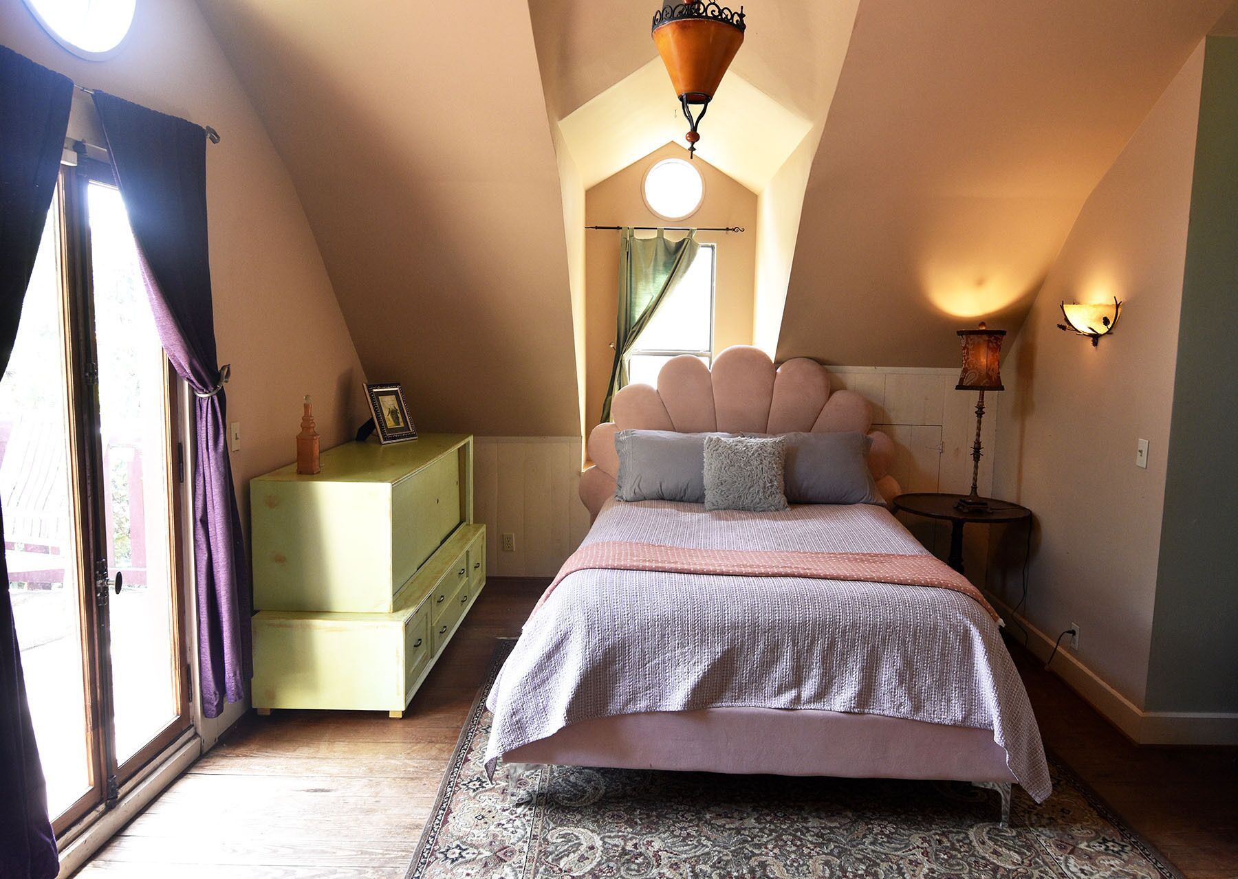 A bedroom with a sloped ceiling, a bed with a scalloped headboard, a dresser, a nightstand with a lamp, and a window with curtains.