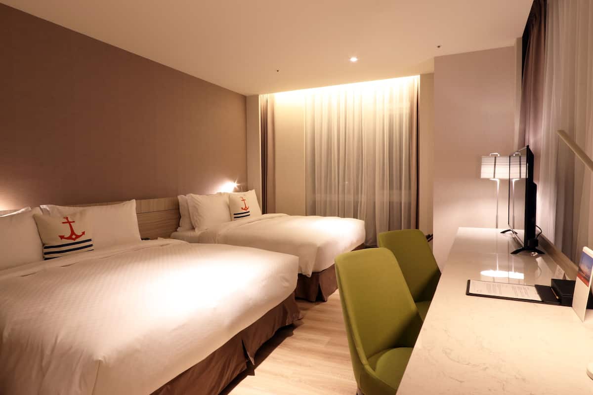 A hotel room with two beds, a desk, a chair, and a lamp.