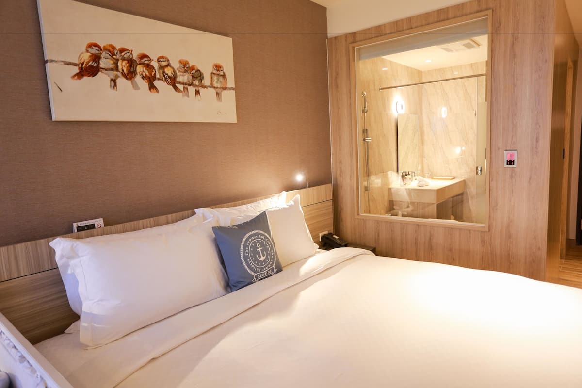 A hotel room with a bed, a painting of birds on the wall, a wooden wardrobe, and a bathroom with a mirror and sink.