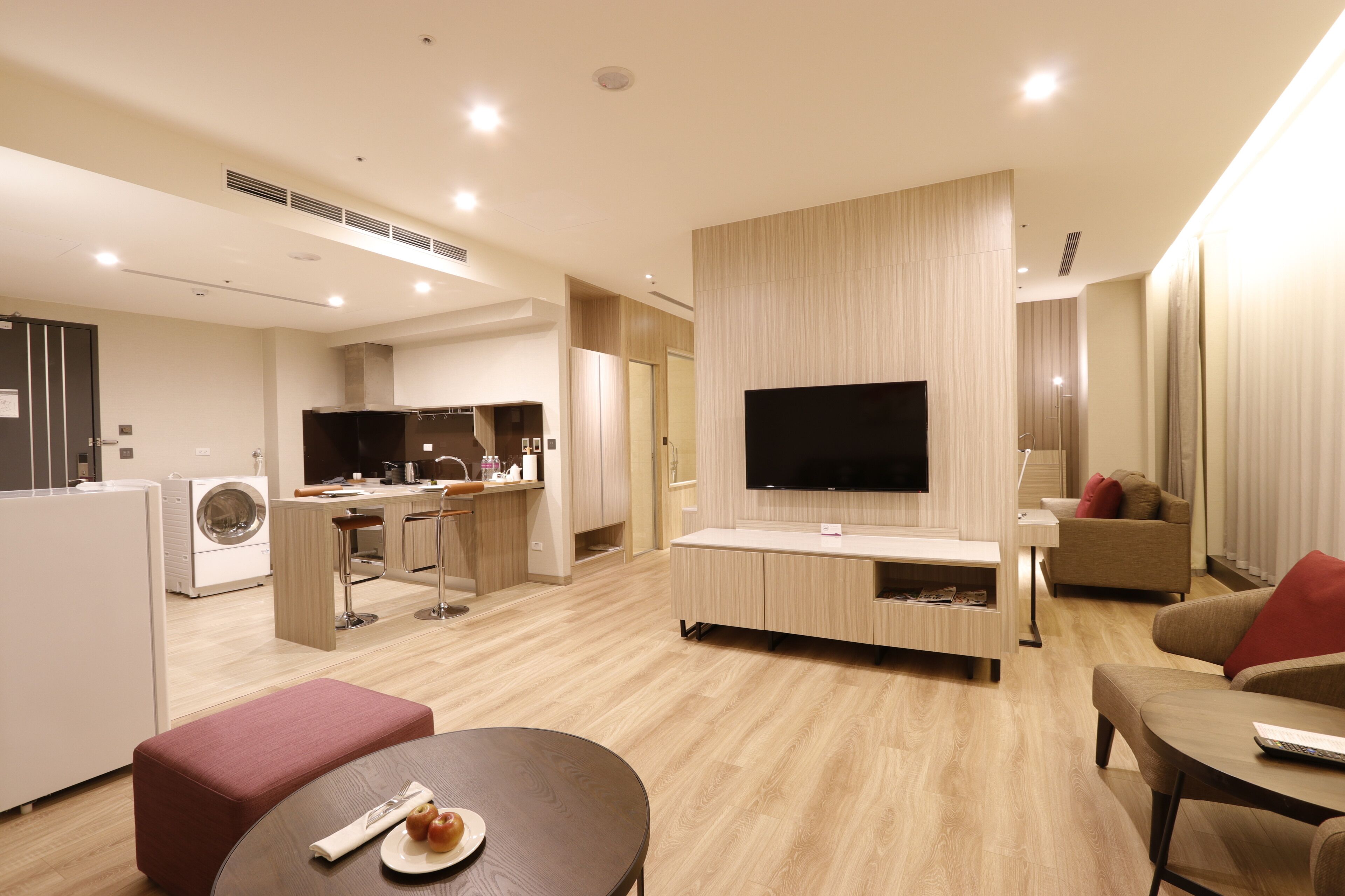 A modern living room with a flat-screen TV, a dining area with a table and chairs, a kitchen area with a washing machine, and a sofa with a red cushion.