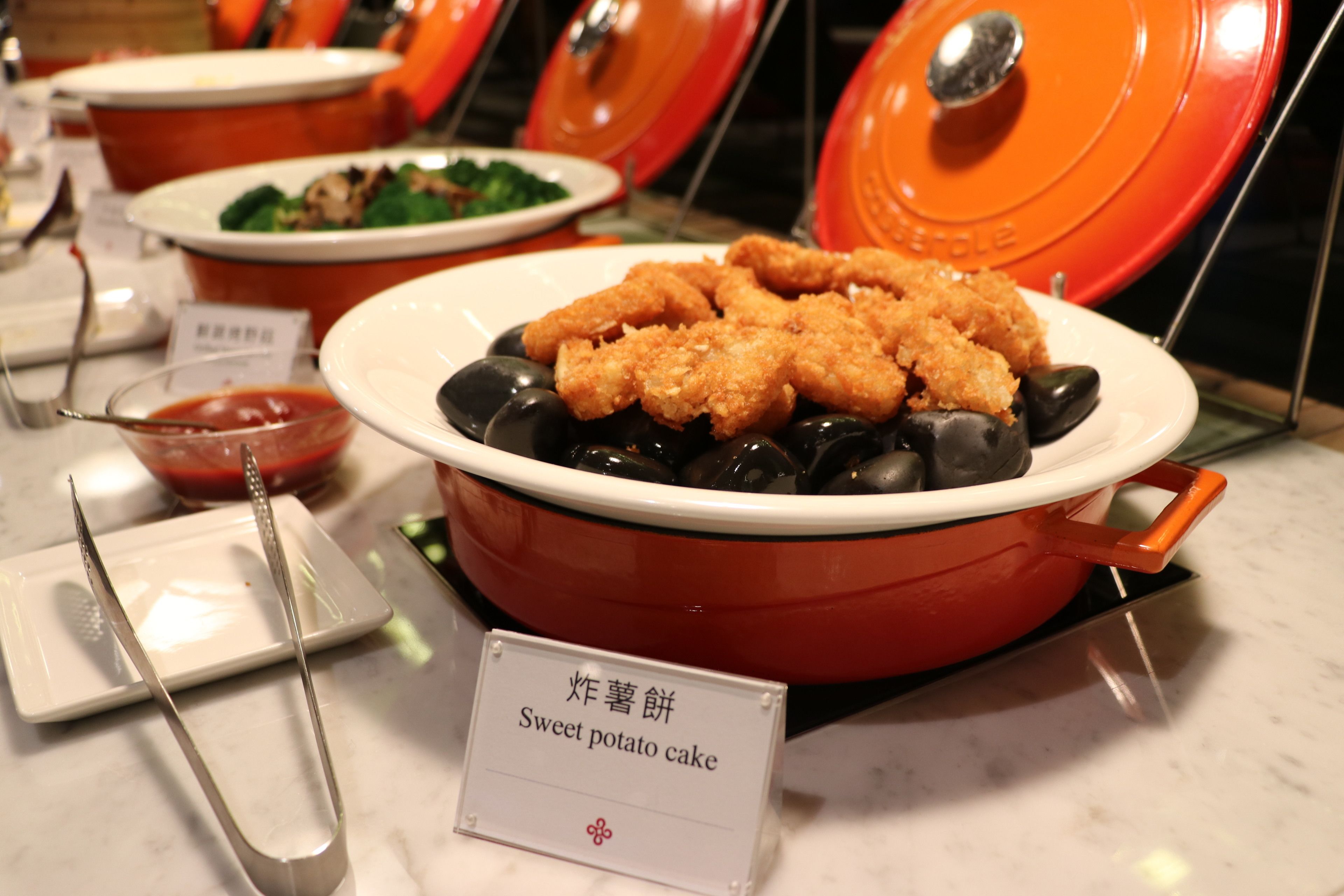 Une table de buffet avec un plat étiqueté « gâteau de patates douces », servi sur un lit d’olives noires.