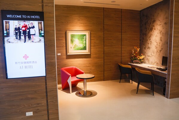 Un moderno vestíbulo de hotel con una pantalla digital de bienvenida, un cuadro enmarcado en la pared, una silla roja, una mesa redonda y un escritorio con un ordenador.