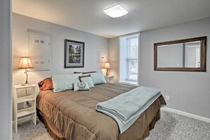 A bedroom with a large bed, two bedside tables with lamps, a window with blinds, a door, and a mirror on the wall.