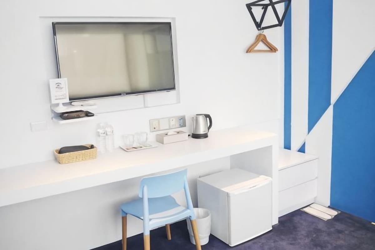 A modern room with a white desk, a blue chair, a television, a kettle, and a small white cabinet.