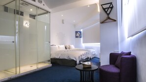 Habitació d'hotel moderna amb un espai de banyera de vidre, un llit amb roba blanca, un butaca morada i una taula rodona petita.