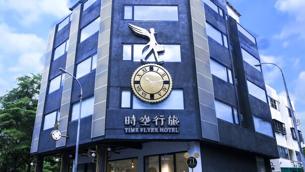 Edifici modern amb un rellotge gran i un logotip d'un cigne, indicat com a "Time Flyer Hotel".