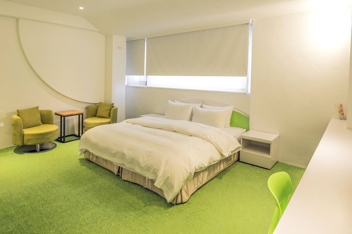 Una habitación de hotel moderna con una cama grande, una alfombra verde, una mesita, una silla y un ventanal con persianas.