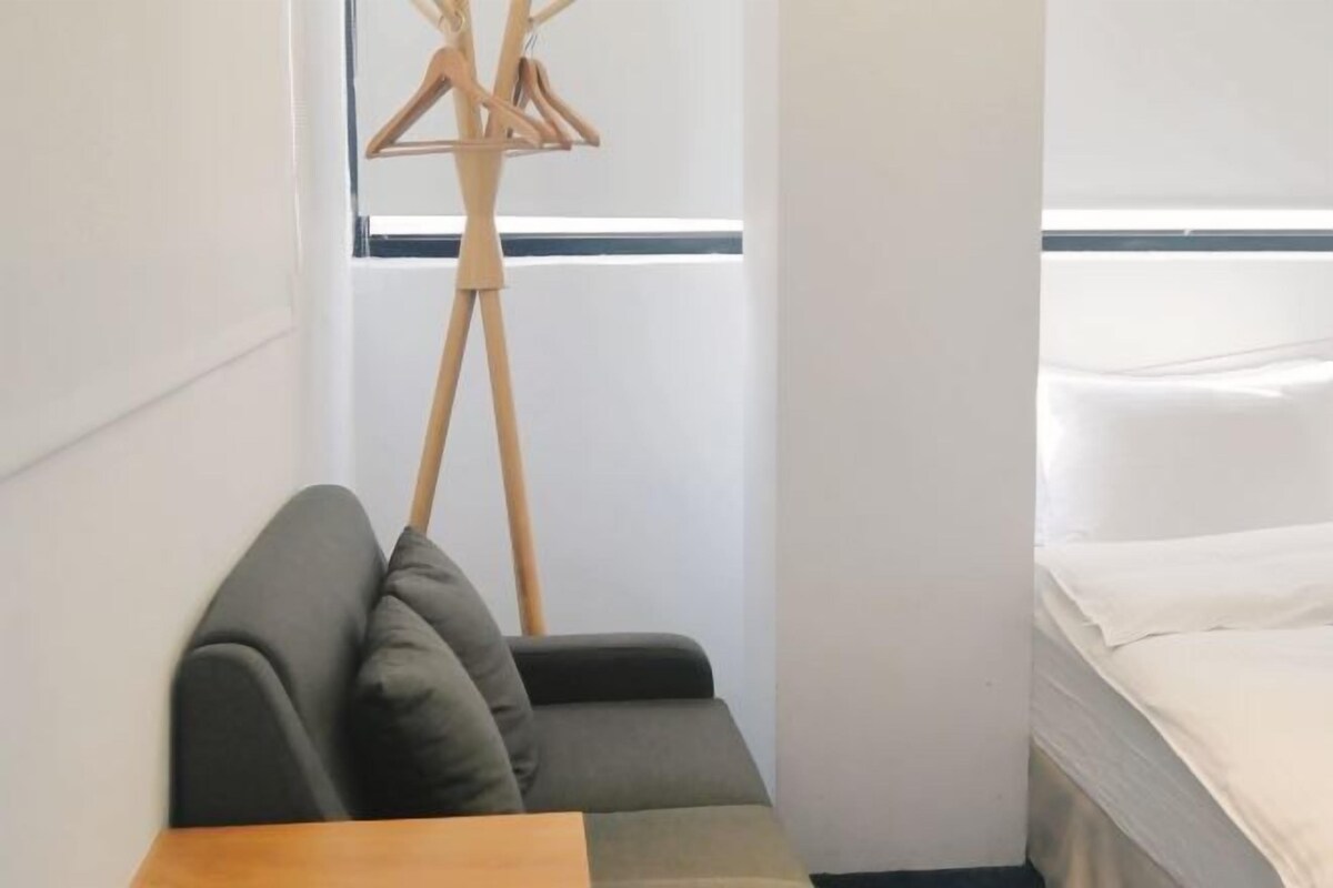 A corner of a room with a wooden table, a grey sofa, and a bed with white bedding.