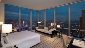 Habitación de hotel moderna con una cama grande, un sofá y un escritorio con vistas a la ciudad de noche.