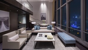 Una sala de estar moderna con sofá, mesa de centro y banco, ventanas amplias con vista a la ciudad y una escalera que lleva a un nivel superior.