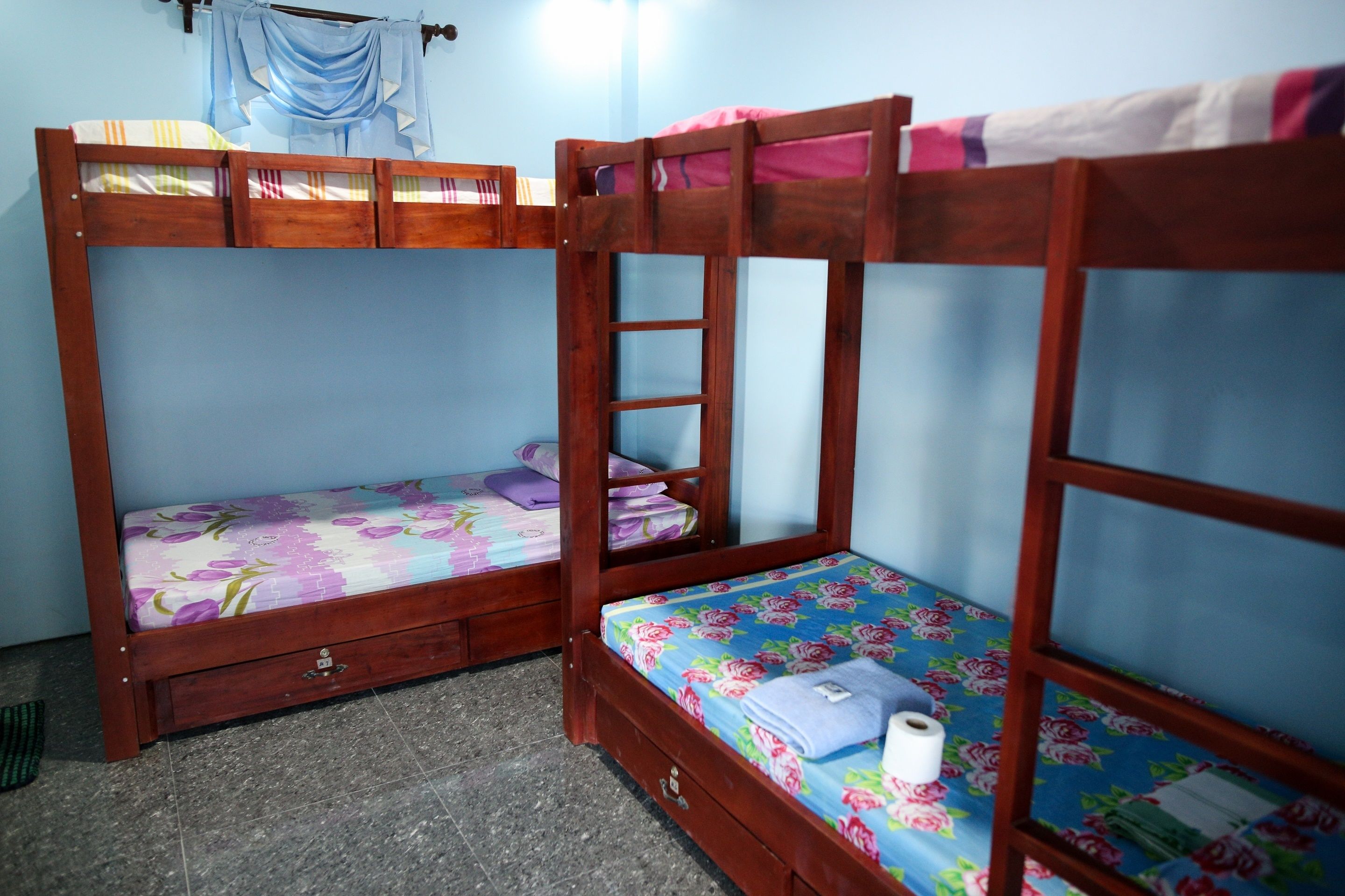 Dos literas con estructura de madera y ropa de cama estampada, un cajón pequeño debajo de la cama inferior y una cortina en la ventana.