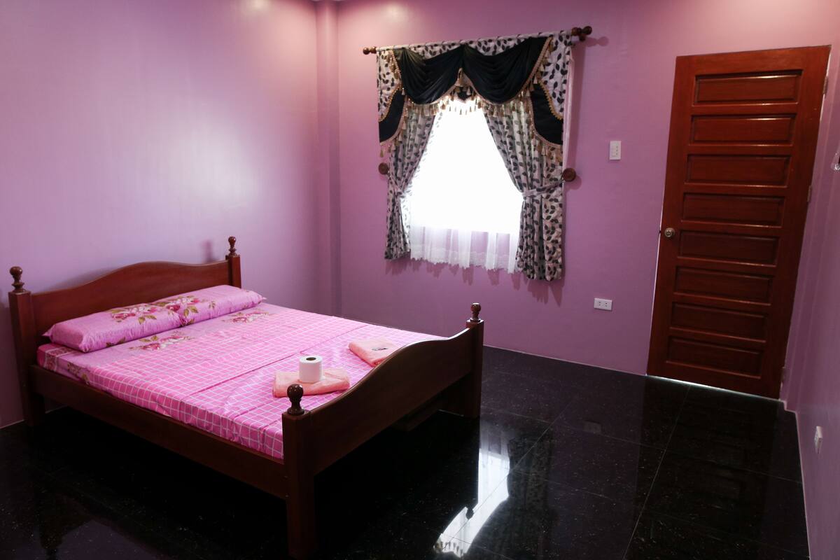 A bedroom with a wooden bed, a window with patterned curtains, and a closed door.