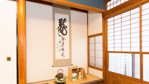 A traditional Japanese room with tatami flooring, a shoji screen, a calligraphy scroll, a tea set, and a small shelf with a book and a teapot. - Guest House Kyorakuya Kinkakuji (Kyoto)