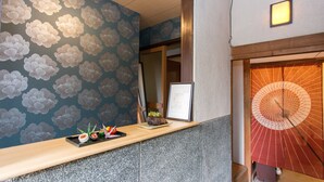 A reception area with a wooden counter, a floral wallpaper, a framed certificate, and a traditional Japanese fan. - Guest House Kyorakuya Kinkakuji (Kyoto)