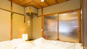 A hotel room with a wooden ceiling, a bed with white bedding, a nightstand with a lamp, and a sliding glass door with a view. - Guest House Kyorakuya Kinkakuji (Kyoto)
