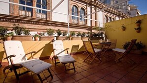 Une terrasse agrémentée de plusieurs chaises longues et de tables, de plantes en pot, et un bâtiment aux fenêtres en arc de cercle en arrière-plan.