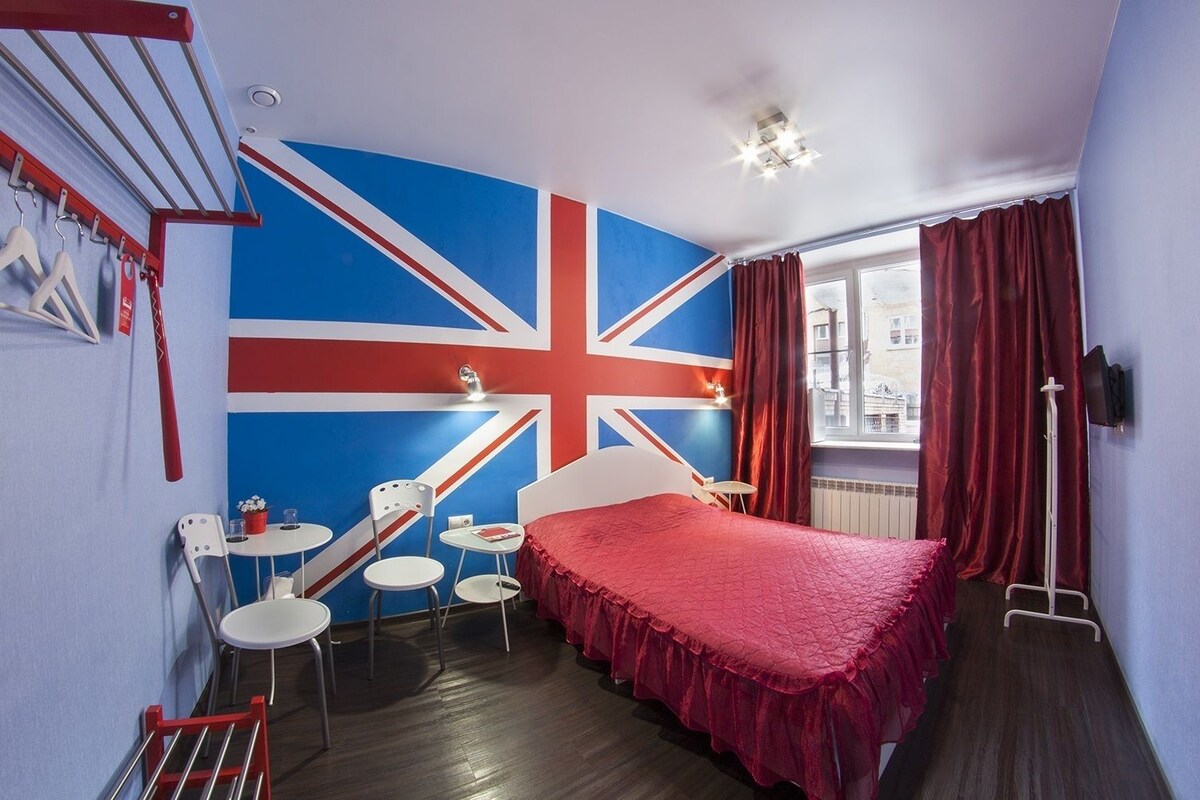 Ein Zimmer mit einer großen Union-Jack-Flagge an der Wand, einem Bett mit roter Bettdecke, einem kleinen Tisch mit Stühlen und einem Fenster mit Vorhängen.