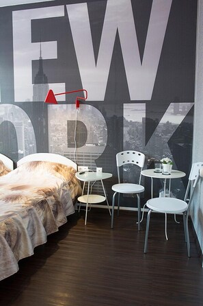 Ein Hotelzimmer mit einem großen Schild mit 'EW', einem Bett mit Daunendecke, zwei weißen Stühlen, einem kleinen Tisch und Blick auf die Stadt.