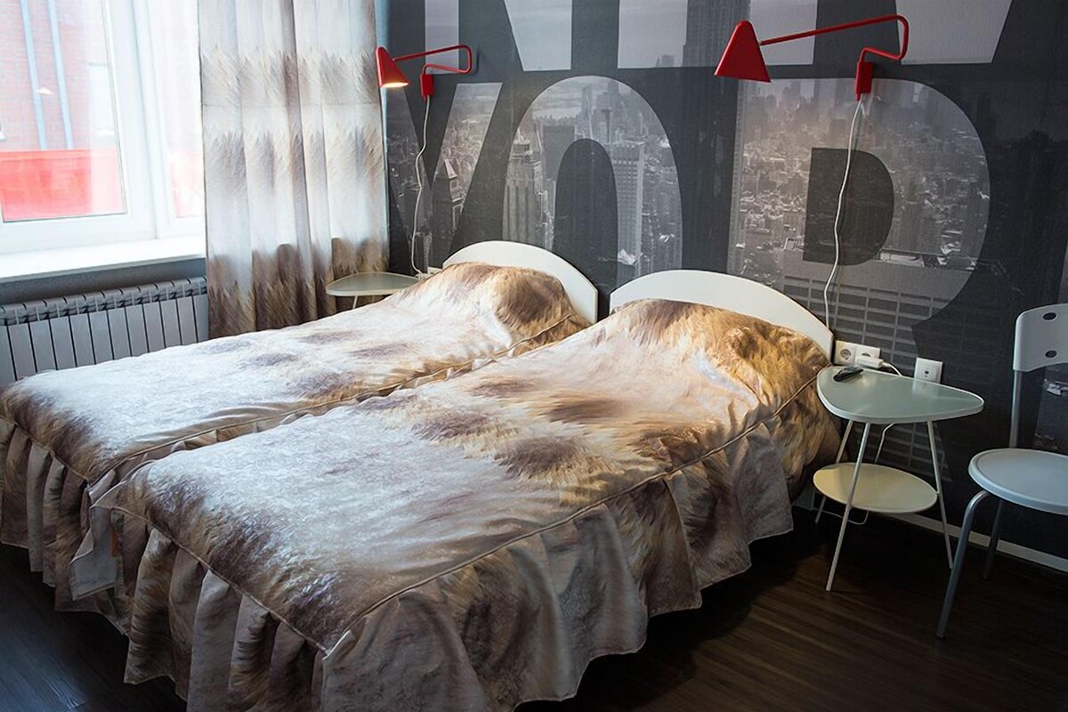 Ein Zimmer mit zwei Betten, einem großen Fenster mit Vorhängen, einer Wandlampe und einem kleinen Tisch.