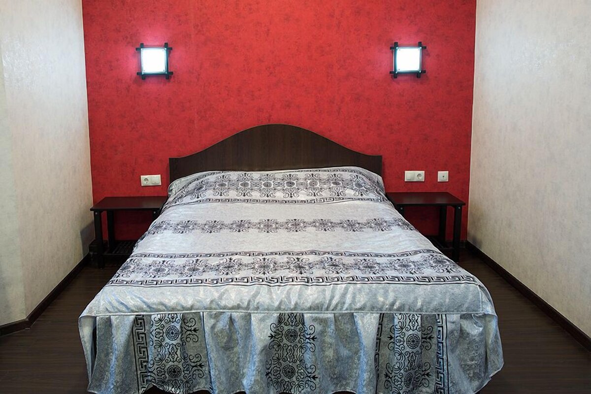 Ein Bett mit gemustertem Überwurf, zwei Nachttische und eine rote Wand mit zwei Wandleuchten.