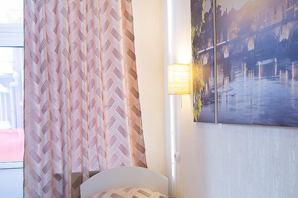 Ein Einzelbett mit einem gemusterten Überwurf, ein Fenster mit Blick auf ein Gebäude und eine Wandleuchte.