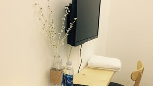 Un bureau en bois avec une télévision, un vase contenant des branches, deux bouteilles d’eau, une télécommande, une bougie et une chaise.