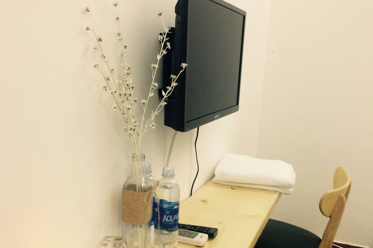 Un bureau en bois avec une télévision, un vase contenant des branches, deux bouteilles d’eau, une télécommande, une bougie et une chaise.