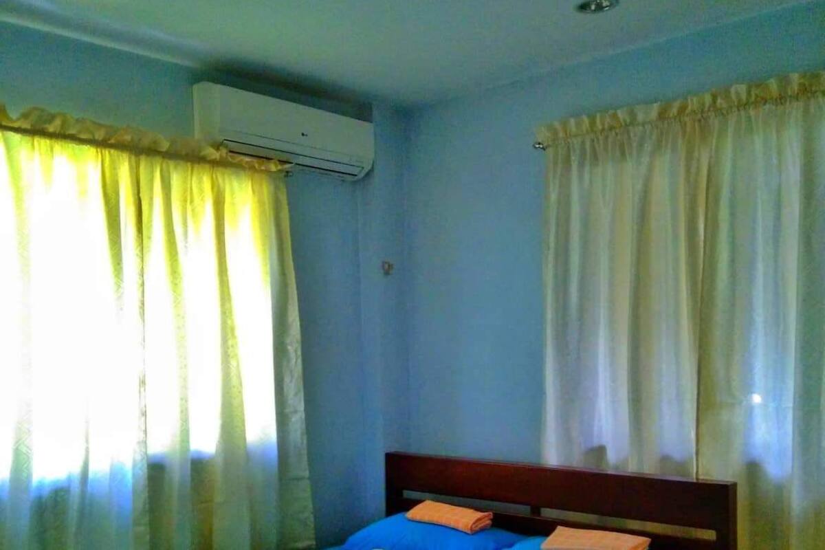 Una cama individual con colchón y almohadas azules, cabecera de madera y ventana con cortinas amarillas.
