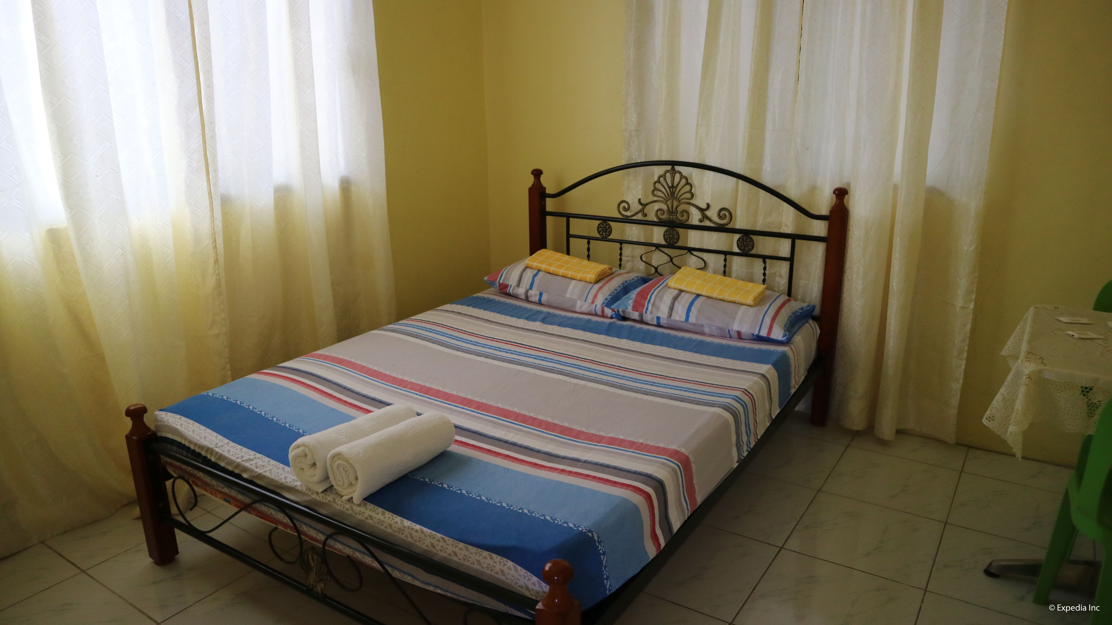 Een enkel bed met een gestreept dekbedovertrek, twee kussens en een opgerolde handdoek. Het bed heeft een houten frame en staat tegen een gele muur.