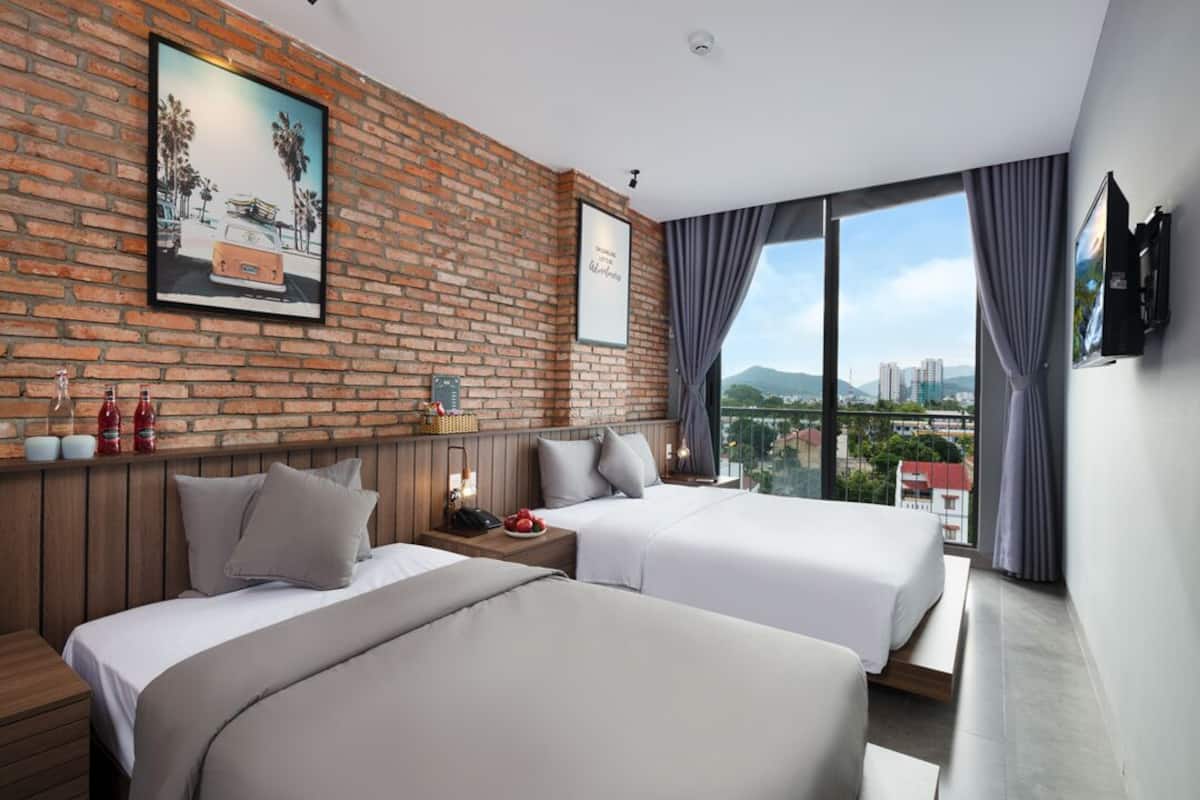Habitación de hotel con dos camas, pared de ladrillo con un cuadro, vista a la ciudad y un televisor en la pared.