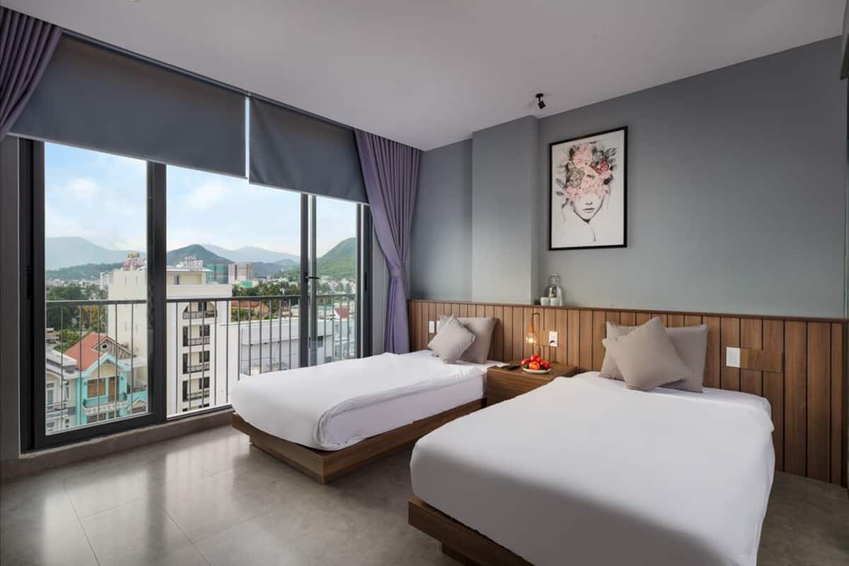 Habitación de hotel con dos camas, un ventanal que ofrece vistas a edificios y montañas, y una obra de arte enmarcada en la pared.