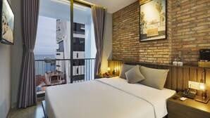 A hotel room with a large bed, a wooden headboard, a nightstand with a lamp, a framed picture on the wall, and a view of buildings through the window. - Marilyn Nha Trang Hotel (Bac Nha Trang)