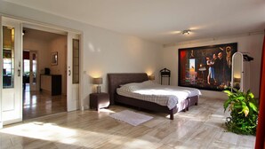 A bedroom with a bed, bedside tables, a large painting, and a potted plant.
