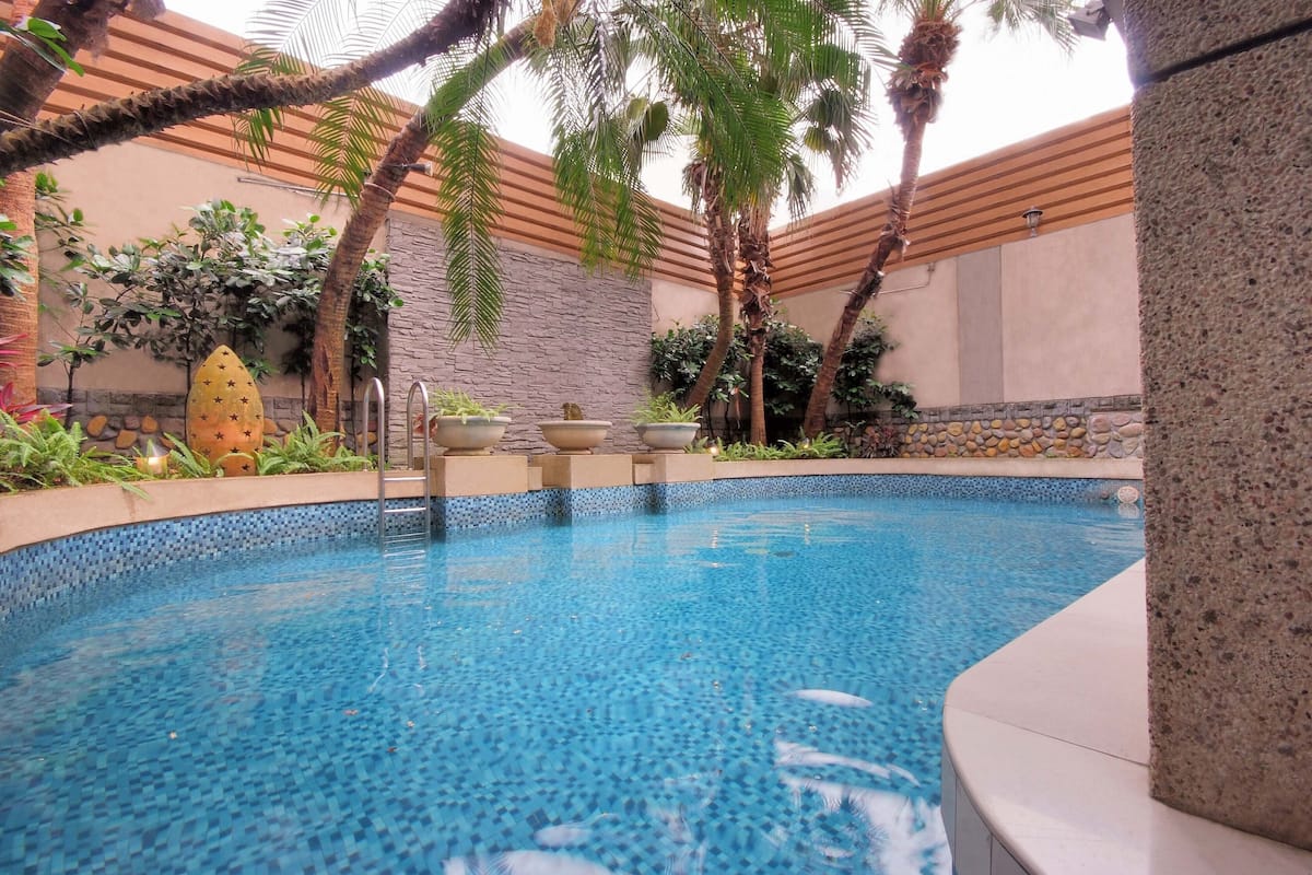 A swimming pool with a tiled edge, surrounded by palm trees and plants, with a stone wall and a wooden ceiling in the background.