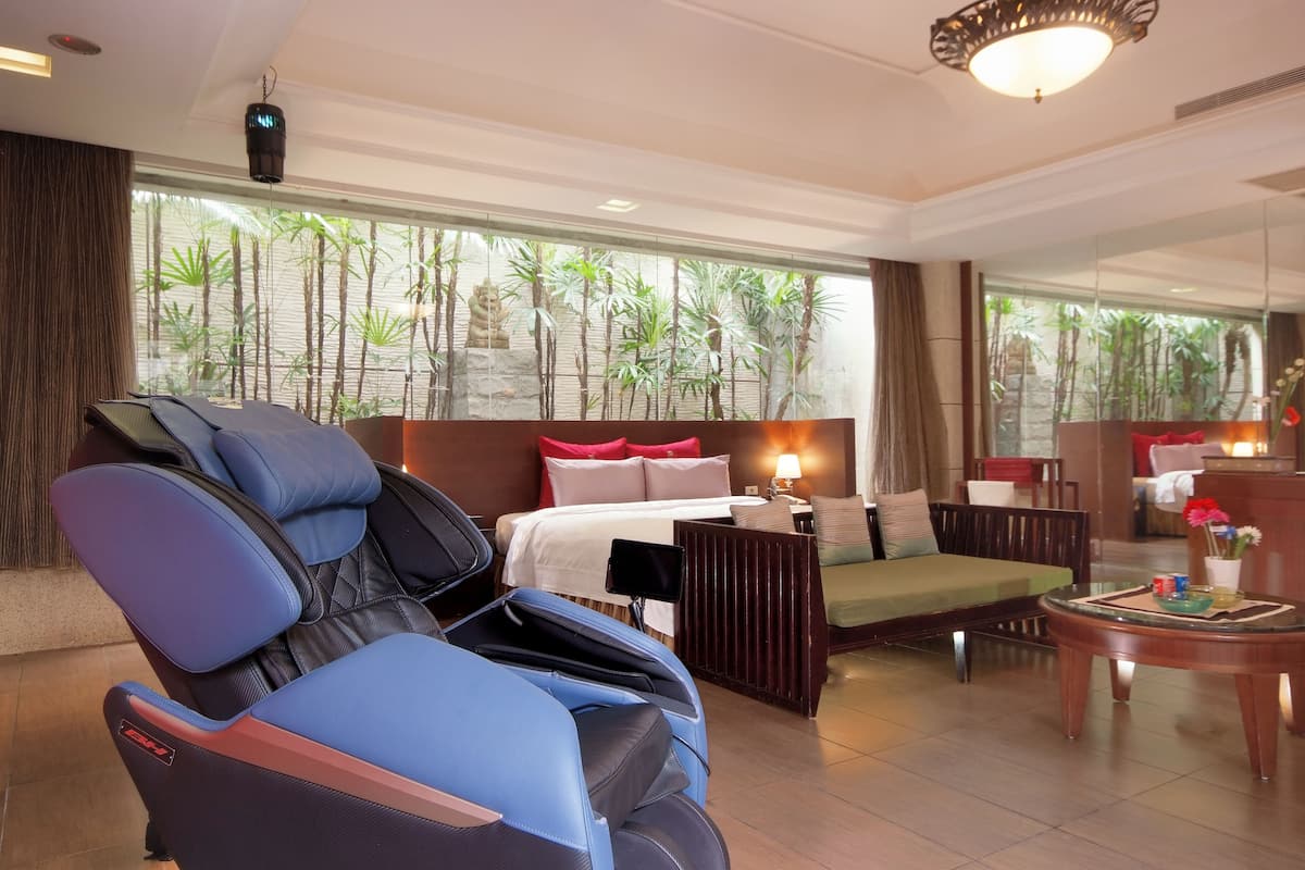 A modern hotel room with a massage chair, a bed with red and white bedding, a sofa, a small table with a vase, and large windows with a view of greenery.