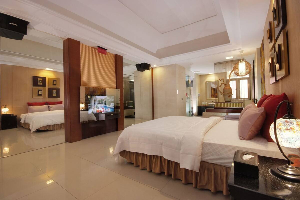 A hotel room with a large bed, a television, a mirror, and a bedside table with a lamp.