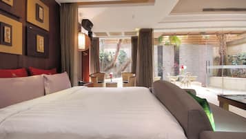 A hotel room with a large bed, a sofa, a dining area with a table and chairs, and a view of the outdoor garden.