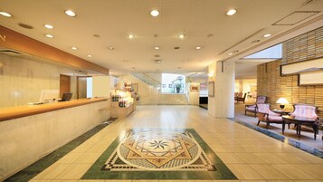Un hall de hotel con una recepción, una escalera que sube a los pisos superiores y un área de descanso con sillas.