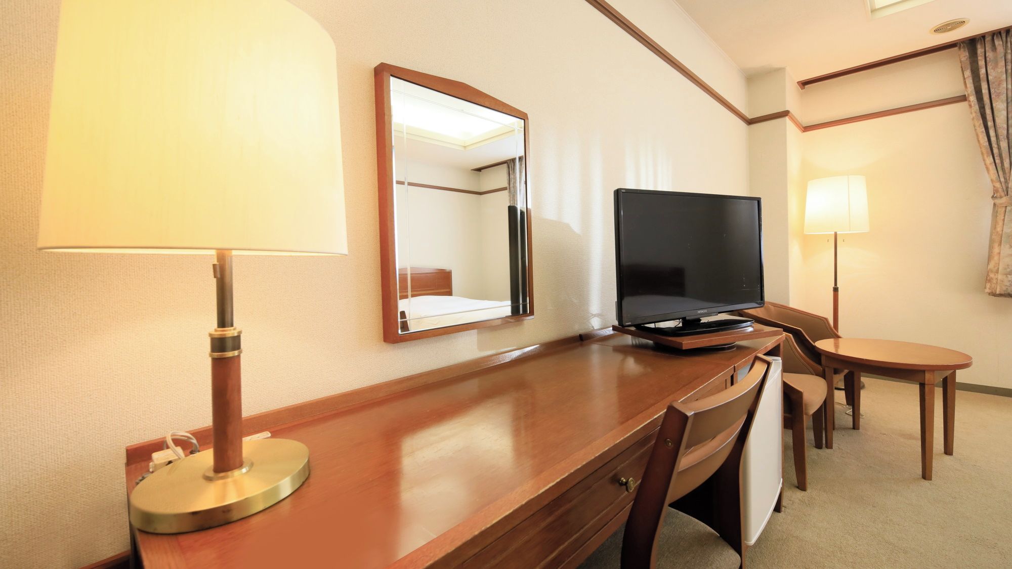 A hotel room with a wooden desk, a television, a mirror, and a lamp.