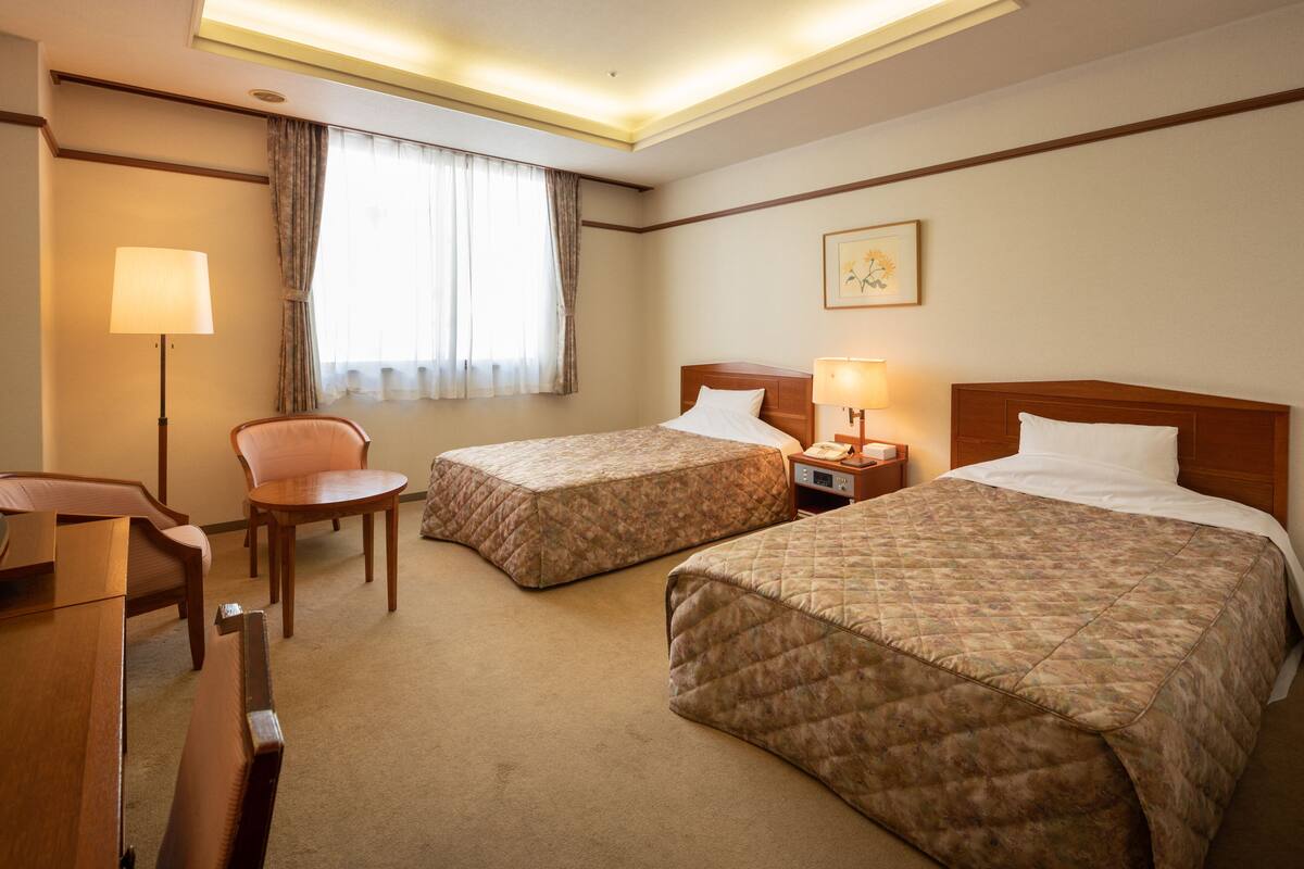 Ein Hotelzimmer mit zwei Betten, einem Schreibtisch, einem Stuhl, einer Lampe und einem fenster mit Vorhängen.