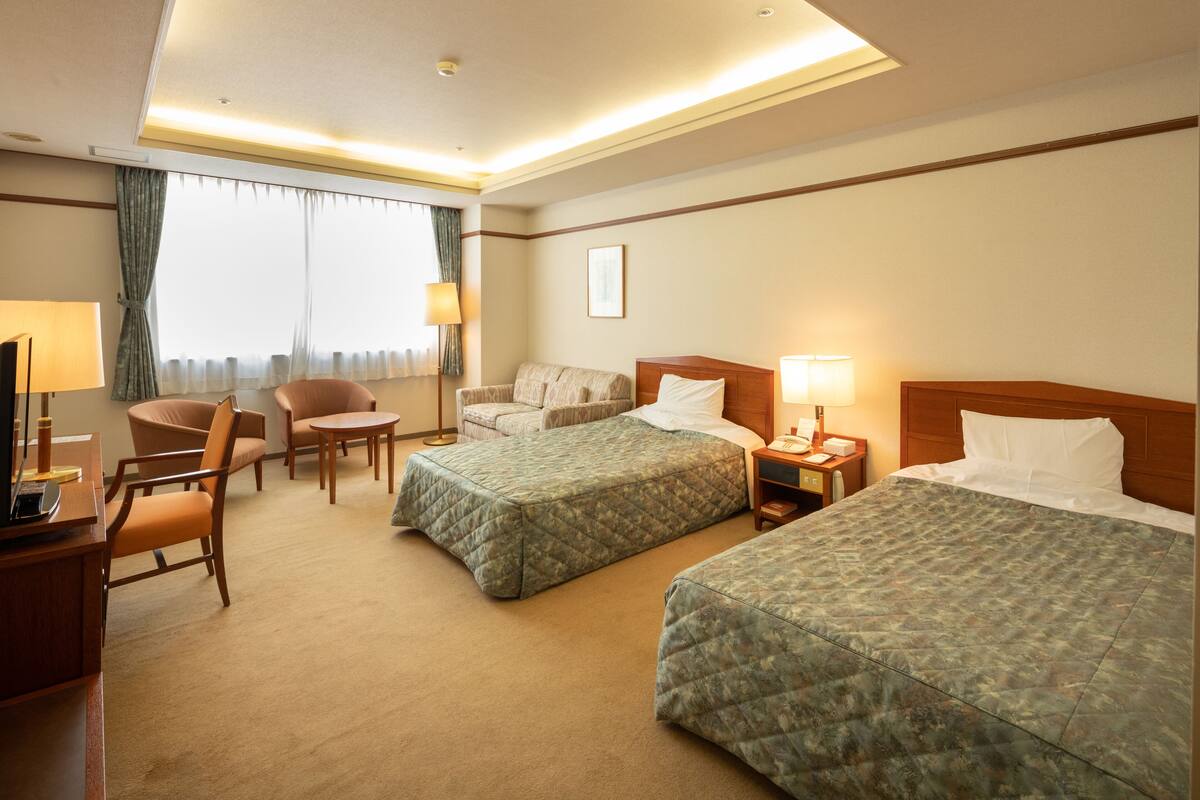 Ein Hotelzimmer mit zwei Betten, einem Schreibtisch mit Lampe, einem Sessel und einem Fenster mit Vorhängen.