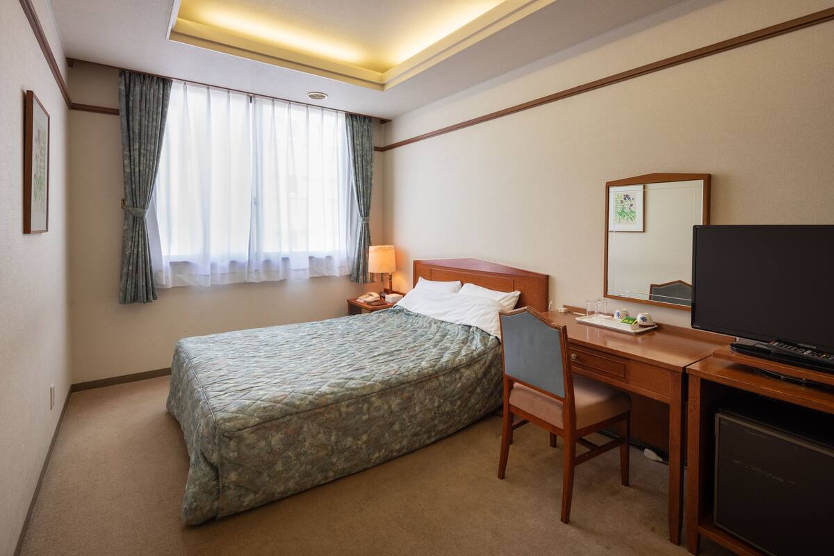 Ein Hotelzimmer mit Bett, Schreibtisch mit Stuhl, Fernseher und einem Fenster mit Vorhängen.