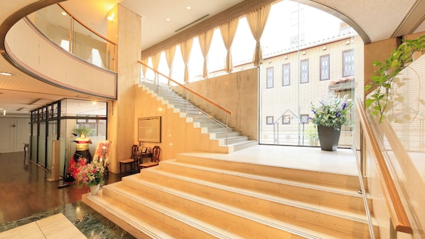 A hotel lobby with a grand staircase, a reception desk, and a view of the building outside.